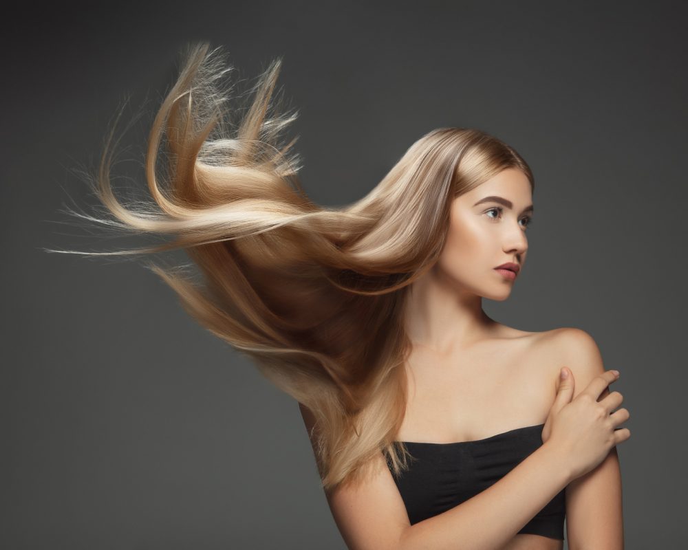 Beautiful model with long smooth, flying blonde hair isolated on dark grey studio background. Young caucasian model with well-kept skin and hair blowing on air. Concept of salon care, beauty, fashion.
